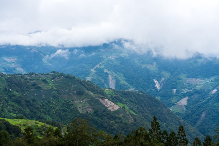 A mountain range with lush greenery, a winding road, and a cloudy sky.