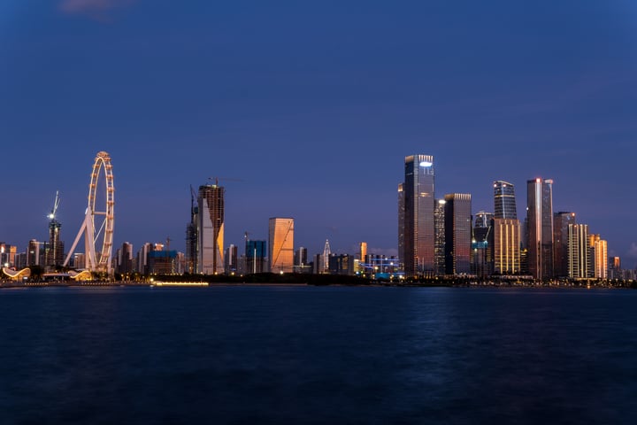 A nighttime view of a city skyline with illuminated skyscrapers, a prominent Ferris wheel, and a calm body of water.