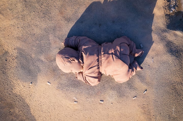 An aerial view of a person lying on the ground, wearing a brown outfit, surrounded by small rocks and a large shadow cast by the person.