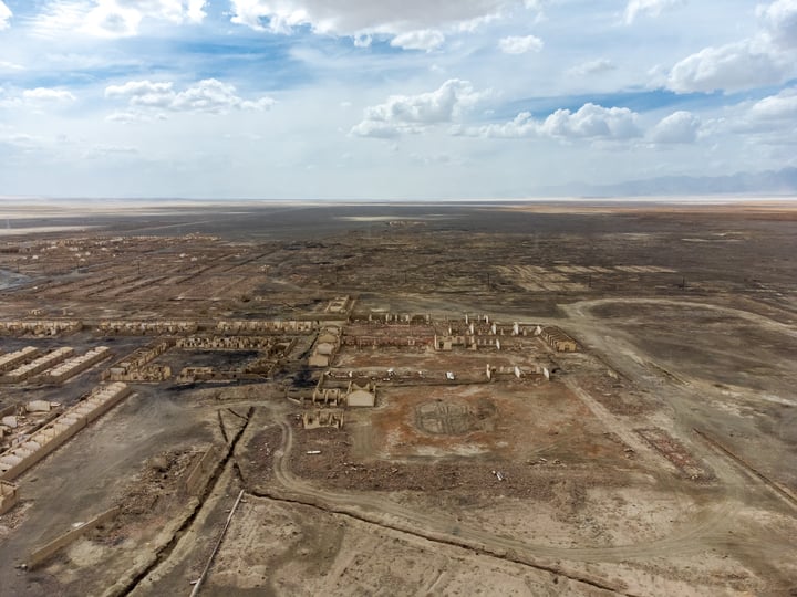 An aerial view of a desert city with a mix of buildings and structures, surrounded by a vast expanse of sandy terrain and a few scattered buildings. The sky is a mix of blue and white clouds, and the city appears to be in a remote location.